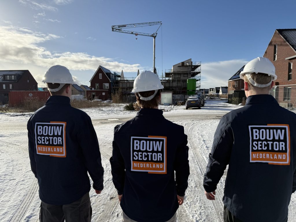 Drie vakmensen van Bouwsector Nederland met witte helm en bedrijfsjas op weg naar een bouwplaats met kraan